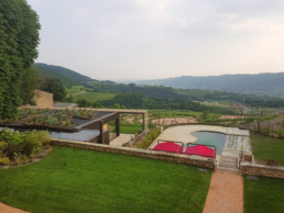 Vista del giardino dal corpo centrale della villa: piscina vista dall'alto, tende da sole rosse, orto circolare e vallata.
